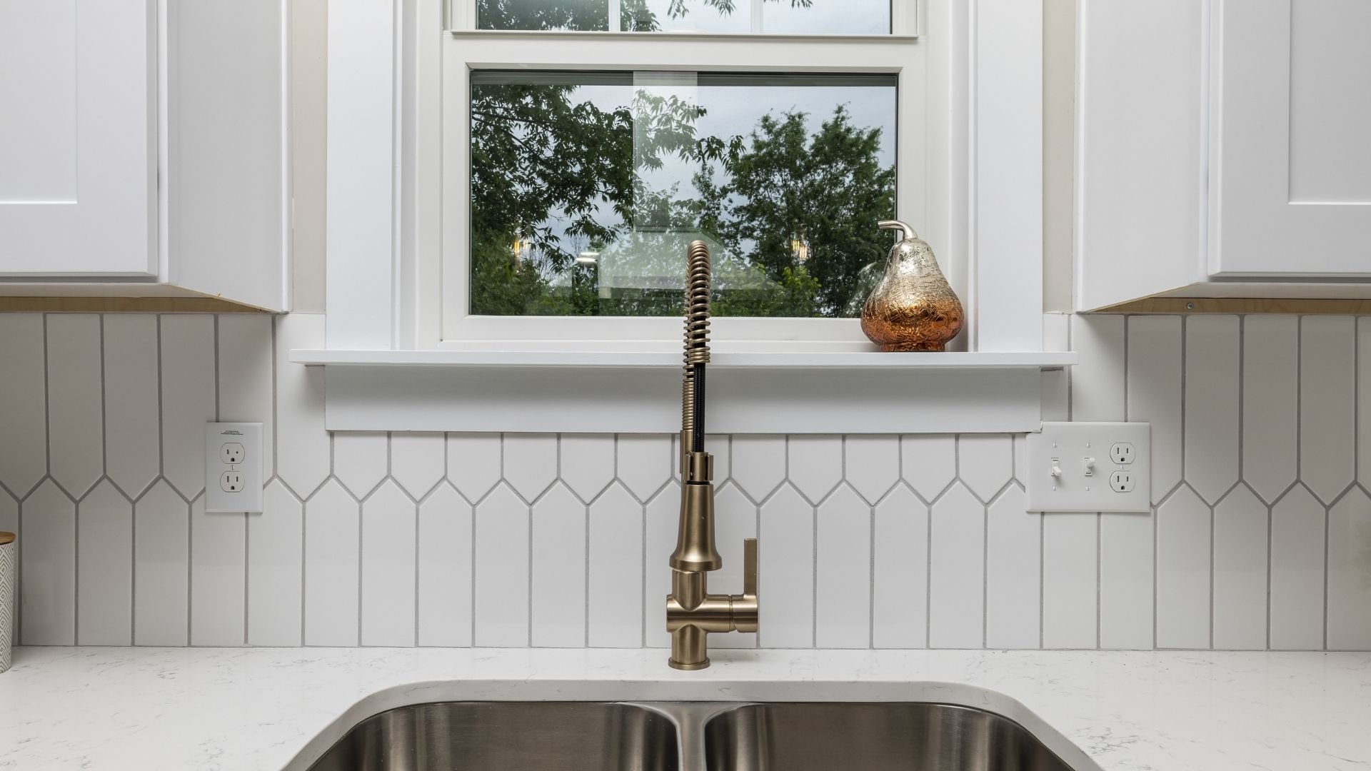 Modern kitchen sink with brass faucet, white tiles, and window view of trees